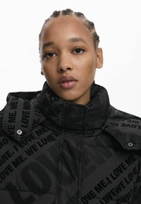 Doudoune noire avec un col haut, ornée de motifs de texte audacieux et répétés. Le tissu a une texture brillante et des détails de couture visibles.