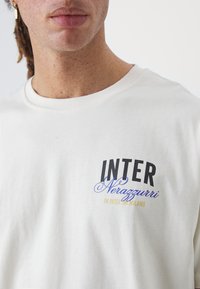 Hvit bomull T-skjorte med teksten "INTER" i svart og "Nerazzurri" i blått, med tilleggstekst i gult, rund hals.