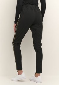 Pantalons slim noirs avec une bande dorée sur le côté, ceinture élastique et deux poches arrière, associés à des baskets blanches. Texture lisse.