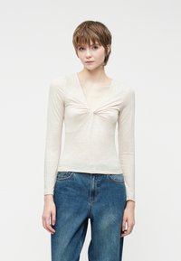 Top de manga larga color beige claro con un profundo escote en V y detalle fruncido en el busto, combinado con jeans azules de corte suelto.