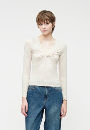 Top de manga larga color beige claro con un profundo escote en V y detalle fruncido en el busto, combinado con jeans azules de corte suelto.