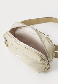 Beige Nylon Tasche mit einem Reißverschluss, verstellbarem Tragegurt und weißem Logo. Das Innere ist mit einer hellen Farbe gefüttert und verfügt über zusätzliche Taschen.