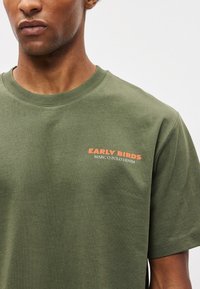 T-shirt de algodão verde oliva com gola redonda. Apresenta as inscrições "EARLY BIRDS" e "MARC O POLO DENIM" impressas em laranja na frente. Mangas curtas.