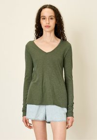 Olivnozeleni dolgi rokav V-izrez majica iz lahkega blaga, z ohlapnim krojem in rahlo teksturo, v kombinaciji s svetlo modrimi jeans kratkimi hlačami.