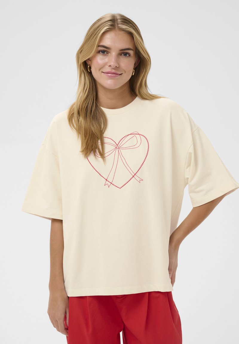 T-shirt beige clair à manches courtes et col rond, avec un motif de cœur rouge et de nœud brodé sur le devant.