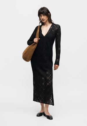 Femme portant une longue robe noire en dentelle avec des motifs floraux, portant un grand sac à épaule marron et des chaussures plates noires.