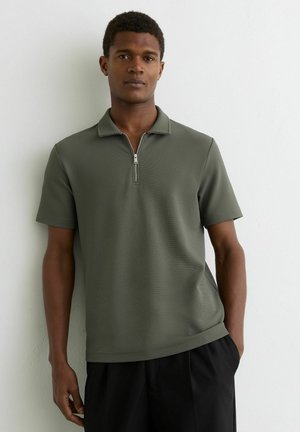 Fiatal férfi rövid göndör hajjal, olívazöld, rövid ujjú, félcipzáras pólóingben, egy egyszerű fehér fal előtt áll.