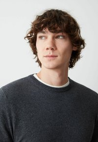 Maglione lavorato a maglia grigio scuro con collo rotondo, indossato sopra una camicia bianca. La texture appare morbida, con una finitura liscia.