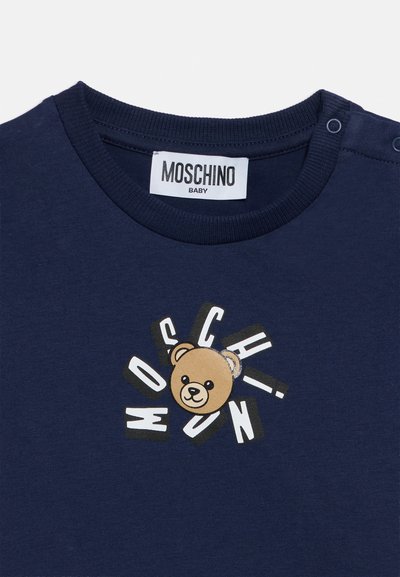 Βαμβακερό παιδικό μπλουζάκι σε ναυτικό μπλε χρώμα με στρογγυλή λαιμόκοψη, διακοσμημένο με γραφικό αρκούδας που περιβάλλεται από το όνομα της μάρκας "Moschino" με έντονα γράμματα.