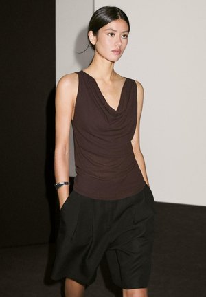 Top in tessuto marrone drappeggiato con scollatura a V profonda, abbinato a pantaloni shorts neri plissettati, su sfondo neutro. Tessuto liscio con una texture morbida.