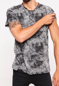Hombre vistiendo una camiseta de manga corta de color blanco y negro con manchas, que muestra un tatuaje de rosa en su brazo superior izquierdo al subirse la manga.