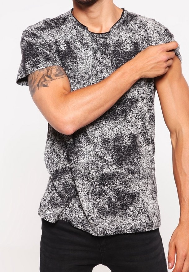 Hombre vistiendo una camiseta de manga corta de color blanco y negro con manchas, que muestra un tatuaje de rosa en su brazo superior izquierdo al subirse la manga.