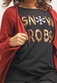 Červený pletený svetr přes černou košili se třpytivým zlatým a stříbrným textem "SNOW PROBS" a sněhovou vločkou, nošený s černými kalhotami.