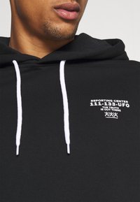 Felpa nera con cappuccio e stringhe bianche, dotata di grafica frontale. Presenta testo stampato: "CENTRO REPORTING 111-133-UFO" e ulteriori elementi di design.