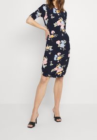 Nainen, joka on pukeutunut tummansiniseen polvipituiseen mekkoon, jossa on pinkkejä, keltaisia ja sinisiä kukkakuosisia printtejä, sekä mustiin avokärkisiin korkokenkiin.