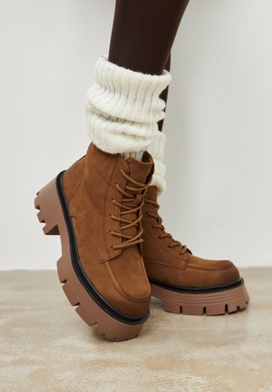 Hnědé semišové kotníkové boty s černými gumovými podrážkami, s designem na šněrování a robustními vroubkovanými podpatky. Nošeny s bílými pletenými návleky na nohy.