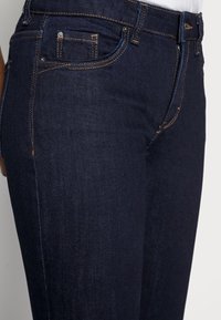 Dark blue jeans made of denim with a slim fit. Features include visible stitching, front pockets, and a zipper fly. Textured finish.