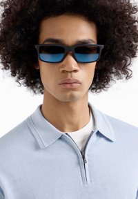 Occhiali da sole neri rettangolari con lenti blu. Indossati da una persona con capelli ricci, abbinati a una polo leggera blu con zip.