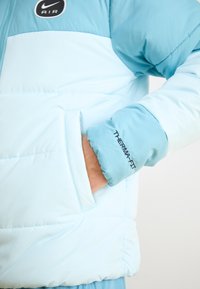 Ljusblå puffjacka med en slät yta, med en svart Nike Air-logotyp, en sidoficka och "Therma-Fit" broderad på ärmslutet.