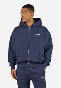 Navy zip-up hoodie with a large hood, front pockets, and ribbed cuffs. Features a white "PEGADOR" logo on the chest and matching joggers.