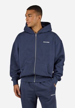 Navy zip-up hoodie with a large hood, front pockets, and ribbed cuffs. Features a white "PEGADOR" logo on the chest and matching joggers.