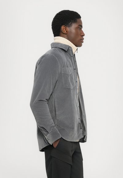 Junger Mann im Profil, der ein graues Cordhemd über einem cremefarbenen Rollkragenpullover und schwarzen Cargo-Hosen trägt, mit Händen in den Taschen.