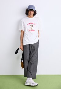 White T-shirt with red text and dog graphic, gray wide-leg trousers, blue cap, New Balance sneakers, holding a small brown bag.