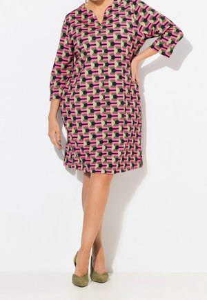 Femme portant une robe mi-longue avec un motif géométrique rose, noir et vert et des talons vert olive, debout, une main sur la hanche.
