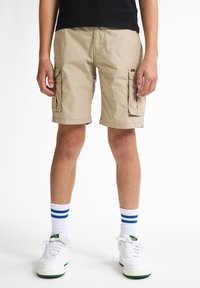 Pantaloni cargo beige con molte tasche laterali, una vestibilità rilassata e una consistenza del tessuto liscia, abbinati a sneaker bianche e calzini a righe.