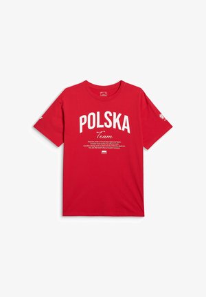 Młody mężczyzna w czerwonej koszulce z białym napisem „POLSKA Team” oraz motywującym przesłaniem wspierającym Reprezentację Polski.