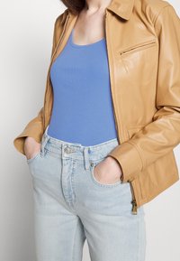 Läderjacka i tan med krage och framficka, som bärs över en blå top. Ljusblå högmidjade jeans med knäppning.