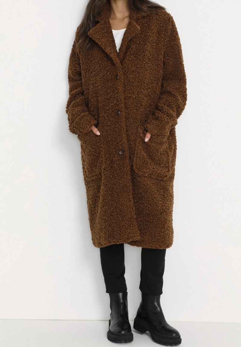 Personne portant un long manteau teddy marron texturé avec poches, pantalon noir et bottines noires, debout devant un fond blanc uni.