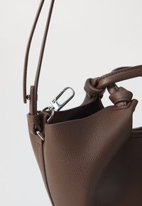 Geantă maro din piele cu o suprafață texturată, având un cârlig metalic, detalii cu mânerele legate și un design cu deschidere în partea de sus.
