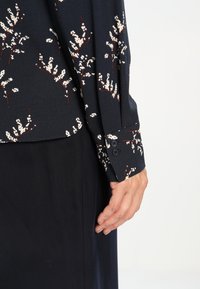 Detailní záběr na ruku odpočívající po boku, oblečenou v tmavě květované dlouhé rukávy blůzy s knoflíkovými manžetami a tmavé sukni.