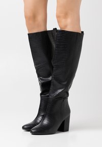 Botas negras hasta la rodilla con textura de cocodrilo, punta redondeada y tacón cuadrado. La superficie es brillante con acentos suaves en la parte superior.