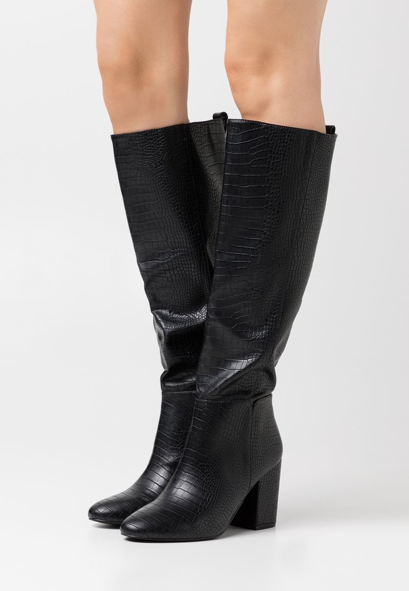 Botas negras hasta la rodilla con textura de cocodrilo, punta redondeada y tacón cuadrado. La superficie es brillante con acentos suaves en la parte superior.
