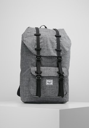 Sac à dos en tissu gris avec des sangles à boucle noire et une poche avant, posé sur un socle blanc sur un fond clair uni.