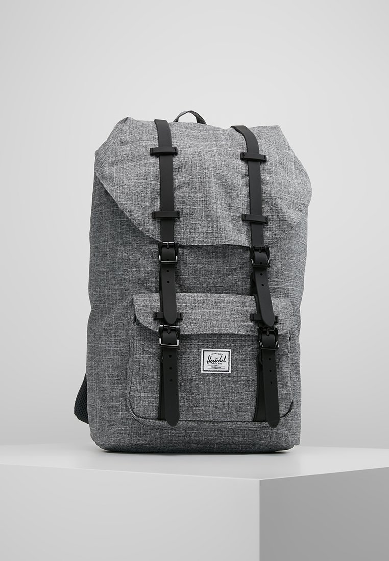 Sac à dos en tissu gris avec des sangles à boucle noire et une poche avant, posé sur un socle blanc sur un fond clair uni.