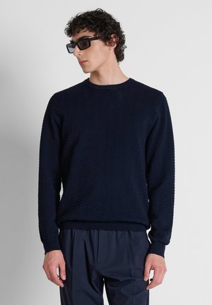 Junger Mann mit lockigem Haar, der schwarze rechteckige Sonnenbrillen, einen strukturierten dunkelblauen Pullover und dunkelblaue Hosen trägt und vor einem schlichten Hintergrund steht.