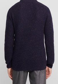 Navyblå stickad tröja med ribbat mönster, långa ärmar och prickig detaljering. Har en hög krage och avslappnad passform.