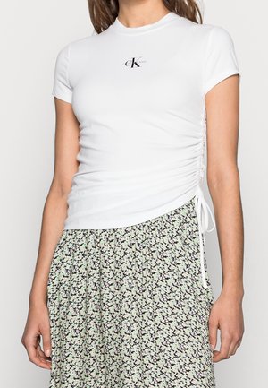 Biały dopasowany krótki top z logo CK Jeans na piersi i marszczeniem po bokach w połączeniu z czarną plisowaną spódnicą w kwiatowy wzór.