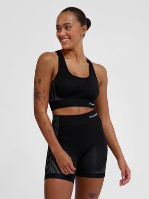 Smilende kvinde med tatoveringer har sort atletisk crop top og højtaljede shorts på mod en ensfarvet grå baggrund.