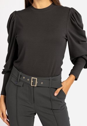 Femme portant un haut noir côtelé à manches bouffantes et un pantalon noir taille haute avec ceinture, une main dans la poche sur un fond uni.