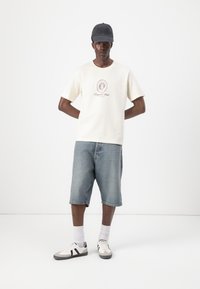 Short-sleeve cream t-shirt with circular red logo, paired with blue denim shorts. Model wears a gray cap and white shoes with black accents.
