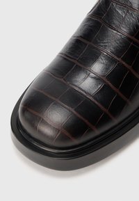 Zapato de cuero negro con textura de cocodrilo, presentando una punta redondeada y una suela de goma negra lisa.