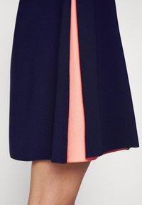 Jupe bleu marine avec un pli central dévoilant une couche intérieure corail, portée au-dessus du genou sur une jambe à carnation claire.