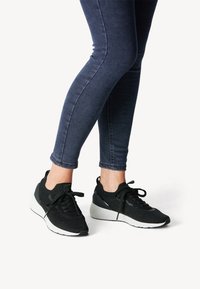 Schwarze sportliche Sneaker mit Mesh-Obermaterial und gepolsterter Ferse, ausgestattet mit einer weißen Sohle und weißen Schnürsenkeln. Kombiniert mit dunkeblauen Skinny Jeans.