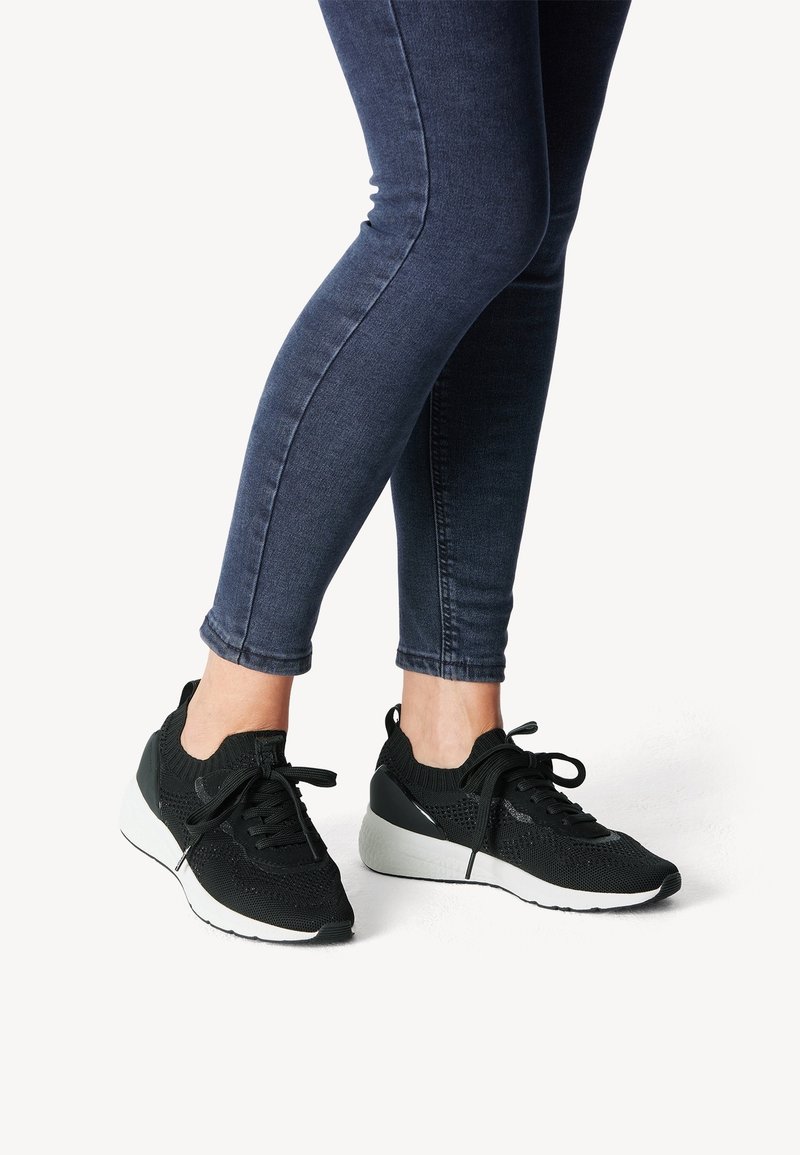 Schwarze sportliche Sneaker mit Mesh-Obermaterial und gepolsterter Ferse, ausgestattet mit einer weißen Sohle und weißen Schnürsenkeln. Kombiniert mit dunkeblauen Skinny Jeans.