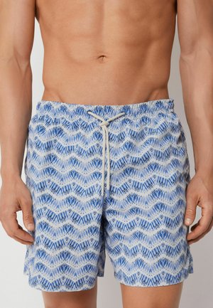 Busto nudo che indossa pantaloncini da bagno blu e bianchi con motivo e coulisse in vita, mani rilassate ai lati su uno sfondo neutro.
