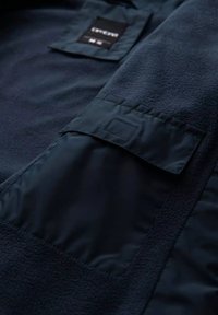 Marineblaue Fleecejacke mit einer glatten Stofftasche mit Klappe und gesticktem Etikett. Der strukturierte Körper kontrastiert mit dem glatten Material der Tasche.